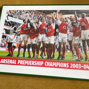 Op de afbeelding: Een ingelijste foto van het Arsenal-voetbalteam dat hun Premier League-overwinning van 2003-04 viert. De spelers dragen rood-witte shirts met het sponsorlogo "O2" op de voorkant. De foto is tegen een groene achtergrond geplaatst en bevat de tekst "Arsenal Premiership Champions 2003-04".