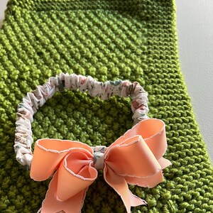 May include: A green knitted baby blanket with a peach-colored bow and a white headband with a floral print.