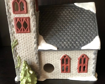 May include: A ceramic church building with a grey roof, white snow accents, and red windows. A small green Christmas tree sits in front of the church.