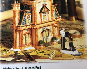May include: A miniature ceramic house with a white and brown exterior, a red roof, and a small telescope on the roof. The house is decorated with windows and a door. The house is sitting on a map with a compass and two figures standing nearby. The text "Admiral's House, Queens Port" is printed below the house.