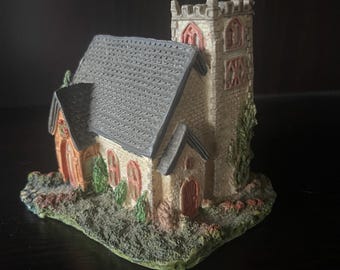 May include: A miniature church figurine with a gray shingled roof, beige walls, and a tall tower. The church has red-framed windows and doors, with green shrubbery and a textured base. The overall design is detailed and decorative.