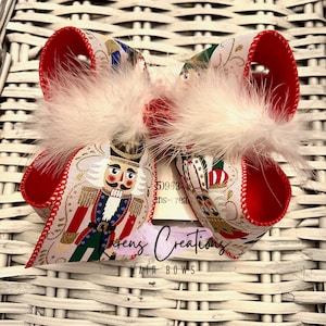 May include: A red and white Christmas hair bow with a nutcracker print and white feathers. The bow is tied with a red ribbon and has the text "Karens Creations Hair Bows" printed on it.