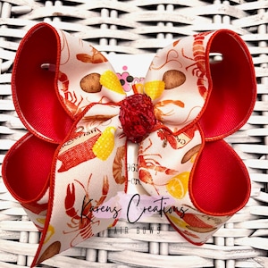 May include: A red and white hair bow with a lobster and seafood print. The bow has a red center with a sparkly red embellishment. The bow is tied with a red ribbon and has the text "Karen's Creations Hair Bows" printed on the bottom.