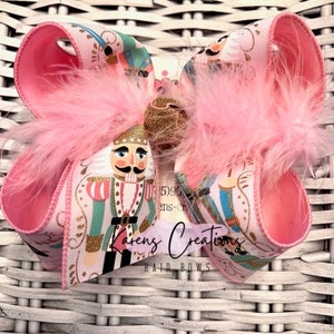 May include: A large pink hair bow with a printed design featuring a nutcracker soldier. The bow has pink feathers and a gold glitter accent in the center. The text "Karen's Creations Hair Bows" is visible.