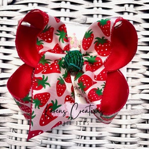 May include: A large, handmade hair bow featuring a vibrant strawberry pattern on a white background, with solid red accents. The bow has a green center and the text "Karen's Creations HAIR BOWS".