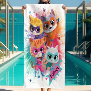 May include: White rectangular beach towel featuring a colorful illustration of four cartoon superhero cats. The cats wear masks and costumes in shades of orange, pink, purple, and blue. The name "Sophia" is written on the towel in a speech bubble.