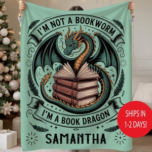 May include: A mint green blanket featuring a dragon illustration with the text "I'M NOT A BOOKWORM I'M A BOOK DRAGON" and the name "SAMANTHA". The dragon is green and black, with a stack of books. The blanket is held up by a person.