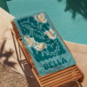 May include: A turquoise beach towel with a white floral pattern and the name "BELLA" in teal lettering.