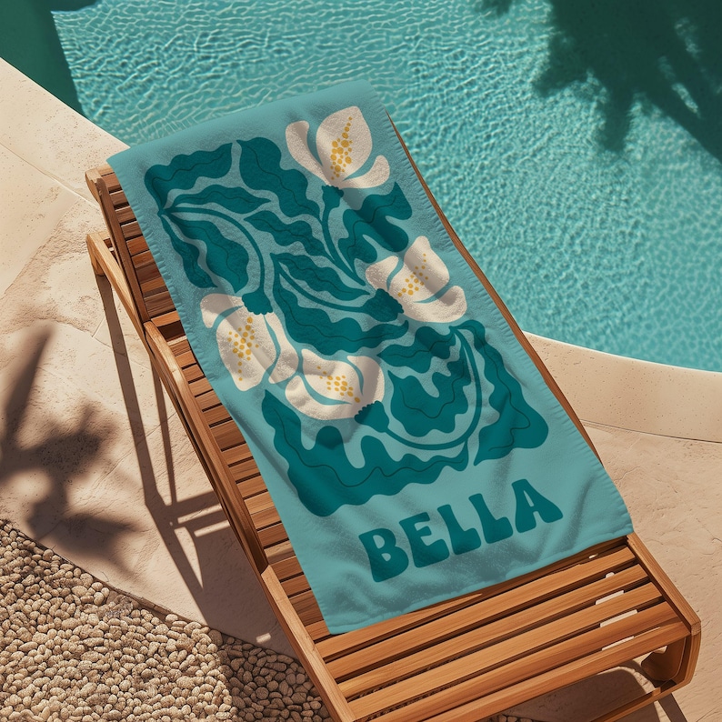 May include: A turquoise beach towel with a white floral pattern and the name "BELLA" in teal lettering.