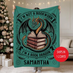 May include: Teal blanket featuring a dragon illustration with the text "I'M NOT A BOOKWORM I'M A BOOK DRAGON" and the name "SAMANTHA". The dragon is green and gold, with a stack of books. The blanket is held up.