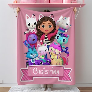May include: A pink fleece blanket featuring a cartoon illustration of a girl surrounded by several cat characters. The blanket has the name "CHRISYINA" on a banner at the bottom. The characters are in various colors.