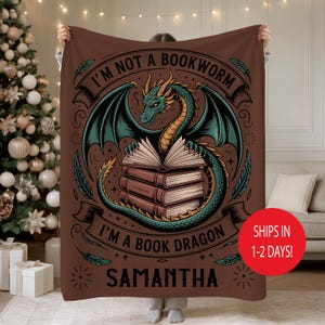 May include: Brown blanket with a dragon design. The dragon is green and teal, wrapped around a stack of books. Text reads "I'M NOT A BOOKWORM I'M A BOOK DRAGON" and "SAMANTHA". The blanket is held up by a person.