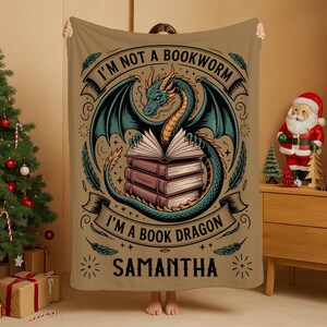 May include: A tan throw blanket featuring a teal dragon with open wings, perched atop a stack of books. The text reads "I'M NOT A BOOKWORM I'M A BOOK DRAGON SAMANTHA". The design includes decorative swirls and starbursts.