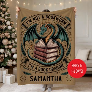 May include: A tan blanket featuring a teal dragon with open wings wrapped around a stack of books. The text reads "I'M NOT A BOOKWORM I'M A BOOK DRAGON" with the name "SAMANTHA" below. The blanket is being held up.