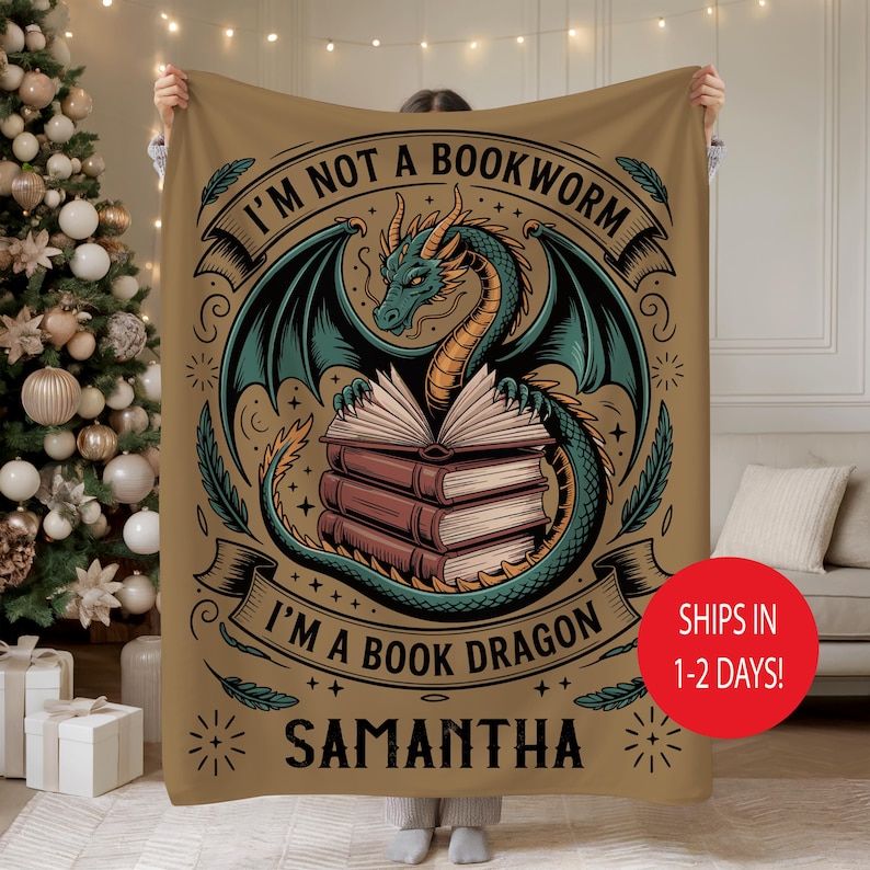 May include: A tan blanket featuring a teal dragon with open wings wrapped around a stack of books. The text reads "I'M NOT A BOOKWORM I'M A BOOK DRAGON" with the name "SAMANTHA" below. The blanket is being held up.