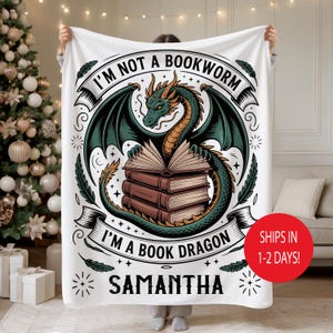 May include: White fleece blanket featuring a green and gold dragon with open books. Text reads "I'M NOT A BOOKWORM I'M A BOOK DRAGON" and the name "SAMANTHA". Decorative elements include stars and flourishes. A red circle indicates shipping in 1-2 days.