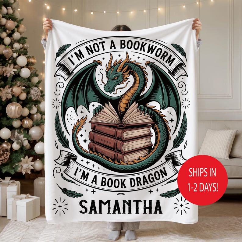 May include: White fleece blanket featuring a green and gold dragon with open books. Text reads "I'M NOT A BOOKWORM I'M A BOOK DRAGON" and the name "SAMANTHA". Decorative elements include stars and flourishes. A red circle indicates shipping in 1-2 days.
