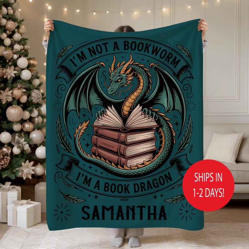 May include: Teal blanket featuring a dragon illustration with the text "I'M NOT A BOOKWORM I'M A BOOK DRAGON" and the name "SAMANTHA". The dragon is green and gold, with wings and is holding books. The blanket is held up by a person.