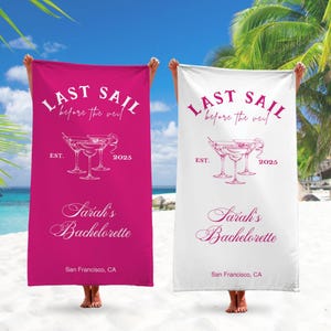 May include: Two pink and white beach towels with the text "Last Sail Before the Veil" and "Sarah's Bachelorette" with a martini glass graphic. The towels are on a beach with a palm tree in the background.