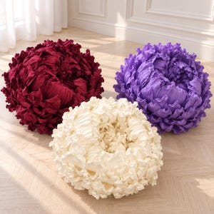 May include: Three round decorative pillows shaped like blooming flowers. One pillow is deep burgundy, one is vibrant purple, and one is cream white. Each pillow features dense, layered fabric petals that create a textured, voluminous, spherical appearance.