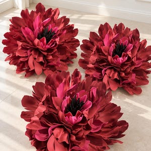 May include: Three decorative pillows shaped like large, blooming flowers. Each pillow features layered, ruffled petals in shades of deep burgundy and vibrant magenta, with a dark black center, creating a realistic, multi-dimensional floral appearance.