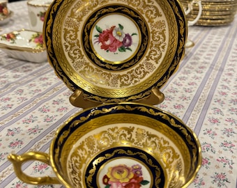 May include: A white teacup and saucer with a gold and blue floral design. The cup and saucer have a scalloped edge and a gold trim.