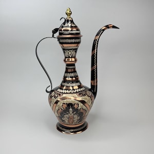May include: An ornate copper and black decorative pitcher with a long, curved spout and a handle. The pitcher features intricate floral and geometric patterns, with a gold-colored finial on the lid. The design is a blend of copper, black, and gold.