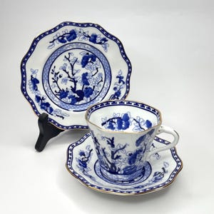 May include: A white teacup, saucer, and plate set with a blue floral design and gold trim. The plate has a scalloped edge and is propped up on a black stand. The teacup and saucer are on a white surface.
