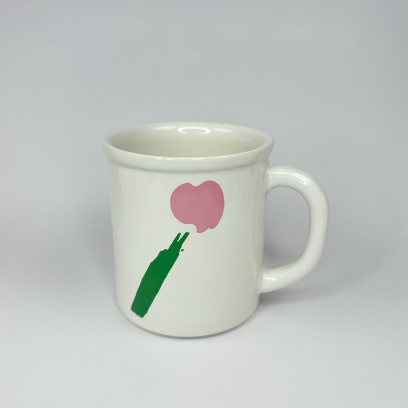 Pode incluir: Caneca de cer&acirc;mica branca com um design abstrato em verde e rosa. O design apresenta uma forma de cora&ccedil;&atilde;o rosa com um caule verde.