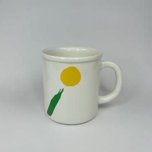 Pode incluir: Caneca de cer&acirc;mica branca com um design abstrato verde e amarelo. O design apresenta um c&iacute;rculo amarelo e uma linha verde.