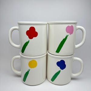 Pode incluir: Conjunto de quatro canecas de cer&acirc;mica branca com desenhos de flores abstratas. Cada caneca apresenta uma flor de cor diferente: vermelho, rosa, amarelo e azul. As flores s&atilde;o pintadas em um estilo simples e moderno com caules verdes.