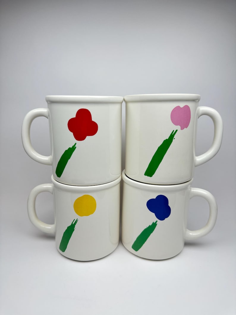 Pode incluir: Conjunto de quatro canecas de cer&acirc;mica branca com desenhos de flores abstratas. Cada caneca apresenta uma flor de cor diferente: vermelho, rosa, amarelo e azul. As flores s&atilde;o pintadas em um estilo simples e moderno com caules verdes.