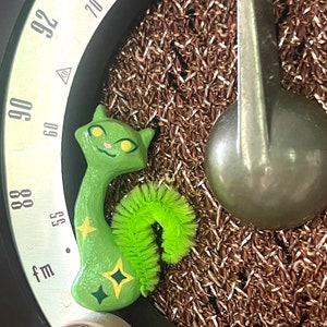 Può includere: Una statuetta di gatto verde con viso giallo e occhi verdi. Il gatto è rannicchiato con una coda verde e gialla. Il gatto è seduto su uno sfondo marrone e argentato con un quadrante che indica "fm" e numeri da 55 a 94.