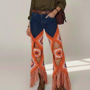 Könnte beinhalten: Dunkelblaue Jeans mit auffälligen orangefarbenen Häkelpaneelen und Fransen. Das Häkeldesign weist geometrische Muster in Orange, Lila und Weiß auf. Die Jeans hat eine hohe Taille und einen braunen Gürtel.