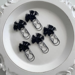 May include: Five black paper clips with a macrame design on top, arranged on a white, decorative plate. The paper clips have a cat-shaped design. The macrame is a dark, solid black color.