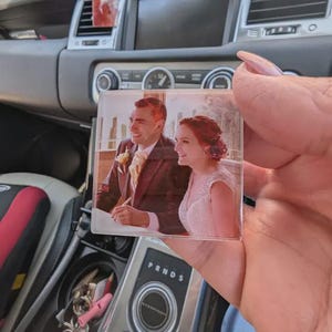 May include: A clear, square acrylic block displaying a wedding photograph of a smiling couple. The man is in a dark suit with a yellow tie, and the woman wears a white wedding dress. The block is held in front of a car's dashboard.