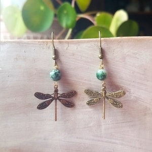 May include: A pair of dragonfly earrings. Each earring features a green and white speckled bead above a detailed bronze-colored dragonfly charm. The earrings have a hook closure.