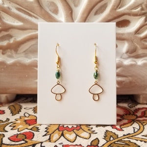 Puede incluir: Pendientes de aro de oro con forma de hongo y cuentas de cristal verde. Los pendientes se muestran en una tarjeta blanca.