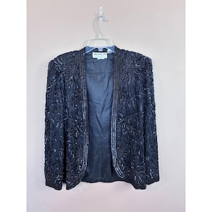 May include: A black sequined jacket with an open front, hanging on a clear hanger. The jacket is covered in intricate floral patterns and has a label that reads "SCALA". The jacket is a formal wear item.
