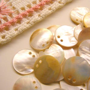 May include: A collection of iridescent, round mother-of-pearl shell beads with three holes, scattered near a section of off-white crocheted lace with pink accents. The beads have a natural, pearlescent sheen.