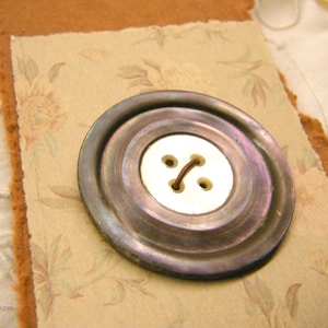 May include: A large, iridescent button with a white center and two brown thread holes. The button is set on a piece of floral patterned paper with a torn, brown edge. The background is a light, neutral color.