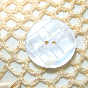 May include: A circular, iridescent white mother-of-pearl button featuring a geometric, recessed square pattern in the center and two small holes for attachment, resting against a pale yellow, open-weave crocheted fabric background.