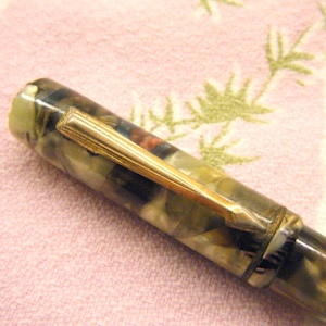 May include: Close-up of a vintage fountain pen with a marbled green, brown, and cream barrel. The pen features a gold-colored clip and trim. The pen is set against a pink background.