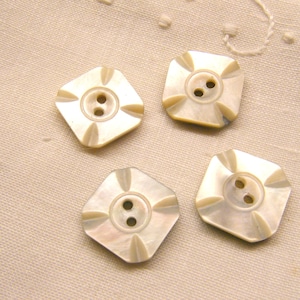 May include: Four off-white, square-shaped buttons with two holes each. The buttons have a decorative design radiating from the center. The buttons are arranged on a light beige fabric.