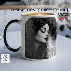 Puede incluir: Una taza de cerámica blanca con un asa negra. La taza presenta una ilustración en blanco y negro de una mujer con los ojos cerrados, sosteniendo un gato. La taza tiene el texto "reveals design once the mug gets warm" impreso en la parte superior. La taza también tiene un logotipo que dice "Cats Books & Bones".