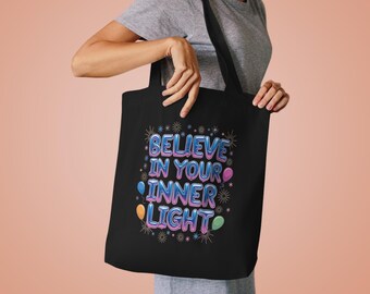 Könnte beinhalten: Schwarze Einkaufstasche mit dem Text "Believe in your inner light" in bunten, ballonartigen Buchstaben. Der Text ist von kleinen Sternen und bunten Ballons umgeben.