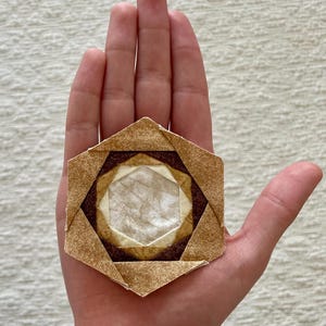 Puede incluir: Una pieza de origami hexagonal hecha de papel marrón y blanco. La pieza tiene un diseño en capas con un hexágono central blanco rodeado de hexágonos marrones.