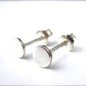 May include: A pair of small, silver stud earrings. Each earring features a flat, circular disc attached to a post with a butterfly backing. The earrings are displayed against a white background, highlighting their simple, minimalist design.