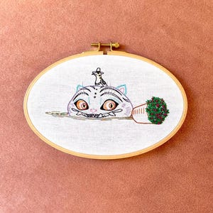 Peut inclure: Broderie ovale représentant un chat fantaisiste avec des yeux orange et un oiseau sur la tête. Un petit pot avec un buisson vert est à droite. Le texte "Velvet Sofa Embroidery" est en bas.