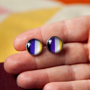Könnte beinhalten: Ein Paar kleine, runde Ohrringe mit einem lila, weißen und gelben Design. Die Ohrringe befinden sich auf einer Hand.